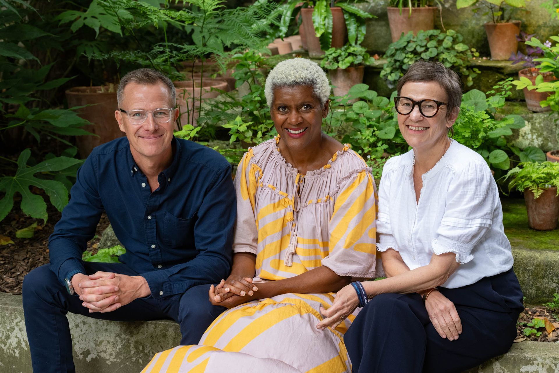 Three people sit together on a stone bench in a leafy garden, smiling at the camera.