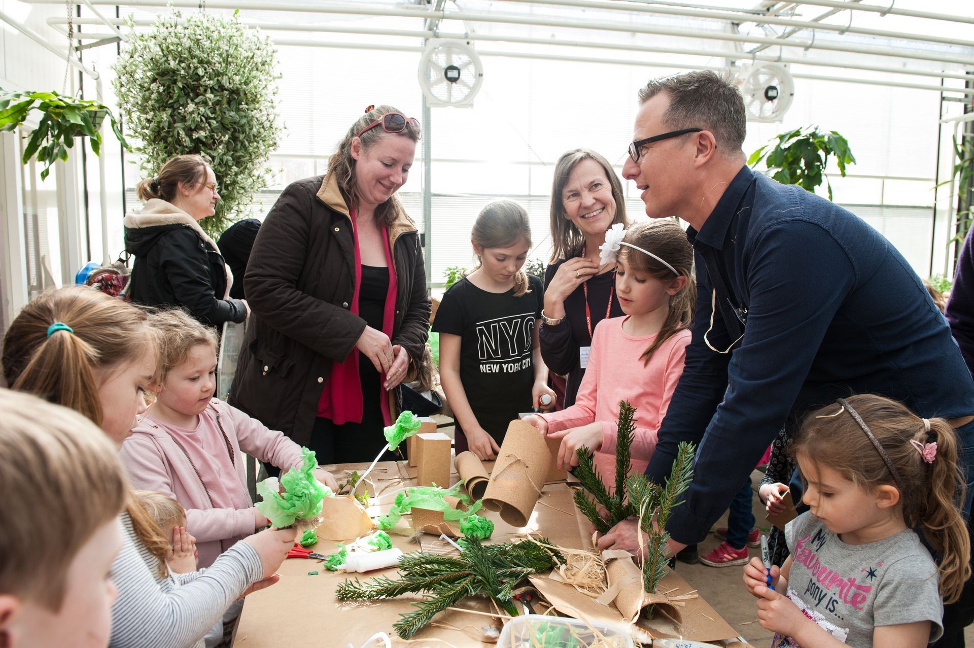 Children and adults craft at a table in a greenhouse, surrounded by plants.