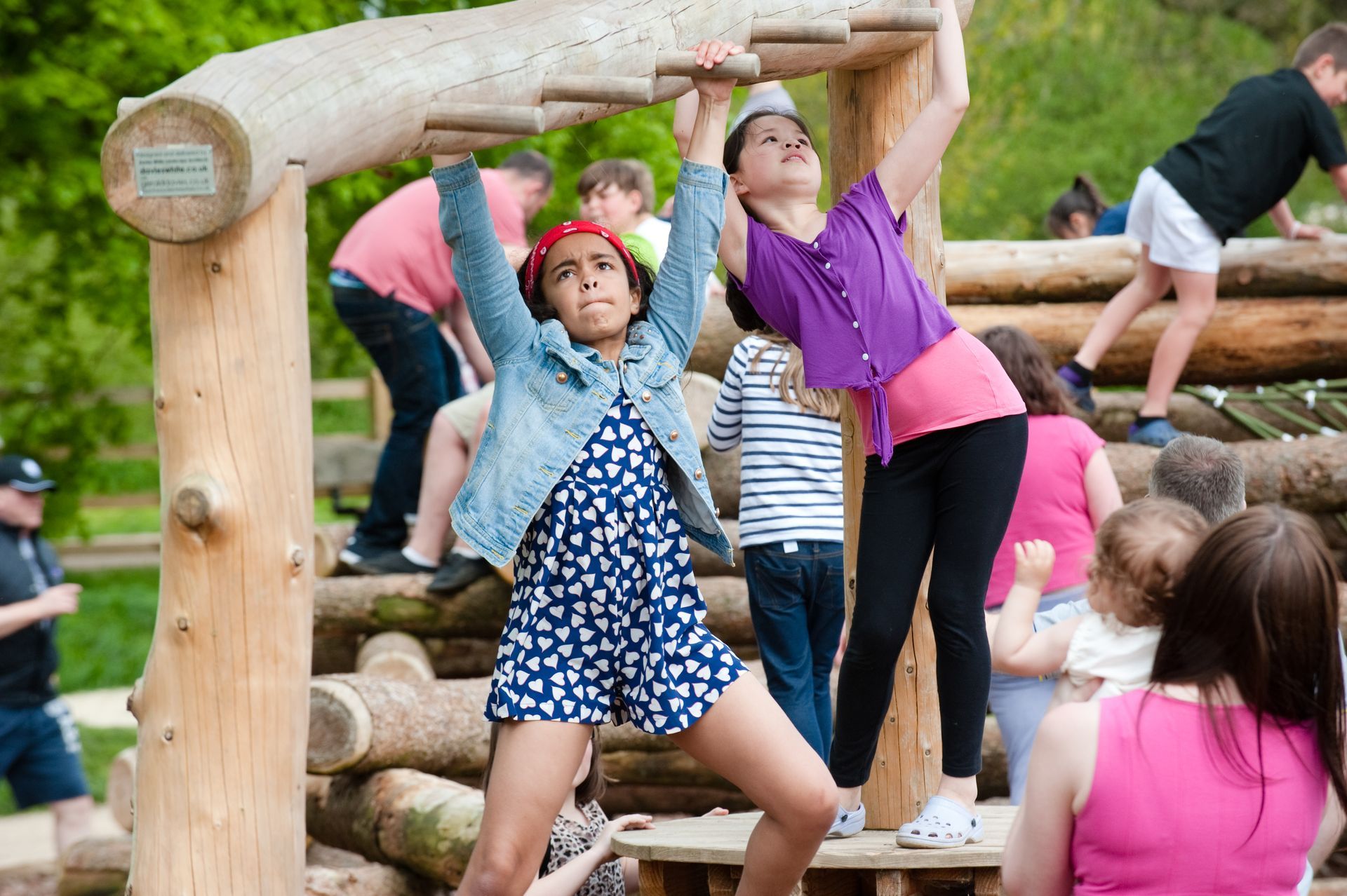 Children playing on a wooden playground structure, one girl hanging, others climbing. Outdoors, sunny.
