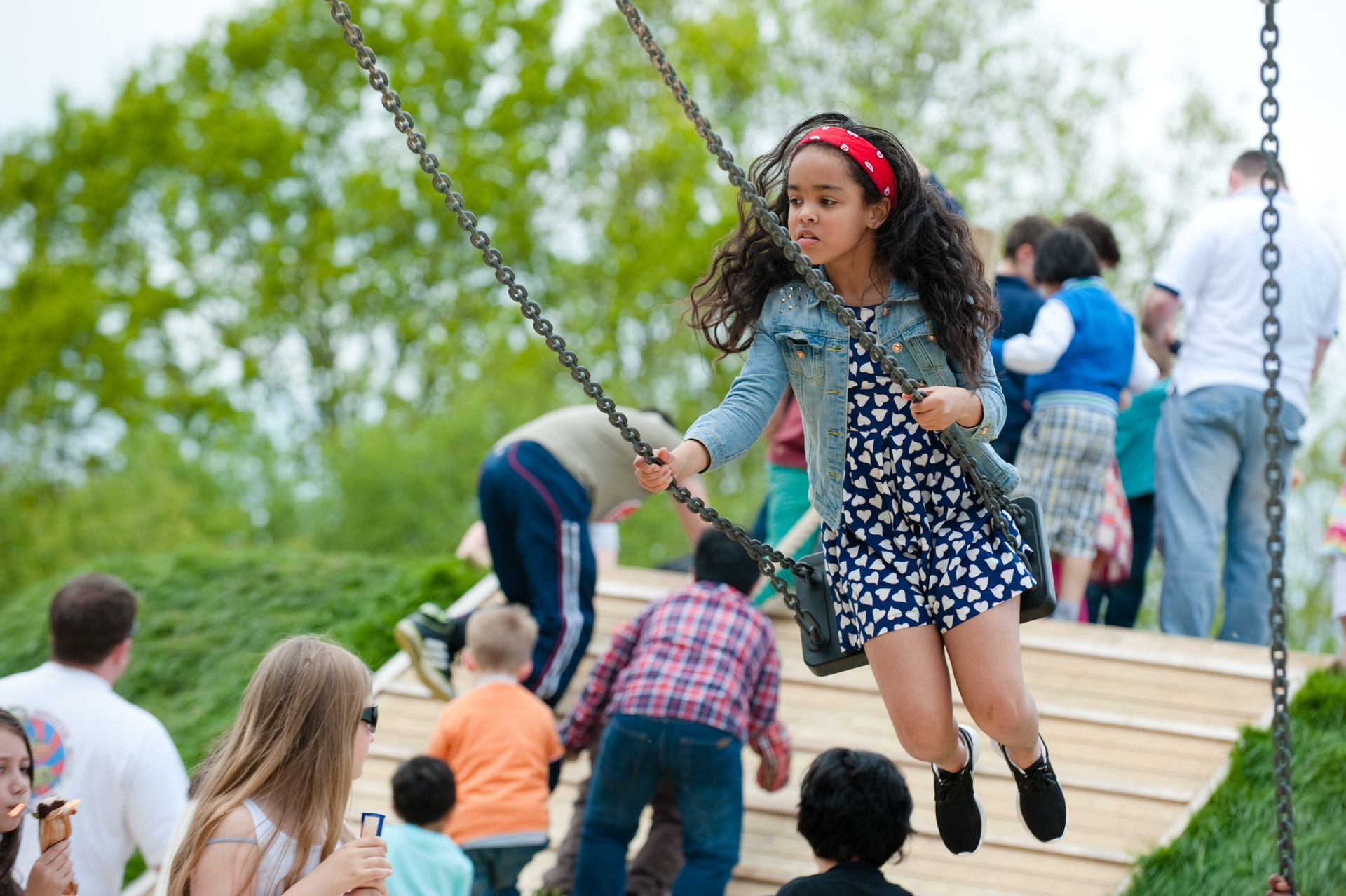 Girl on a swing set at a playground, wearing a blue dress and jean jacket, with other children nearby.
