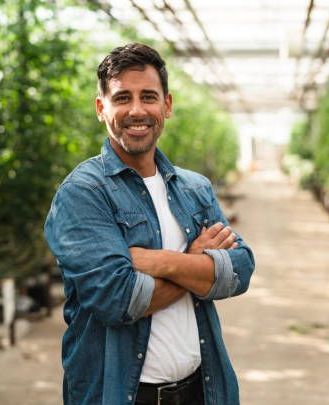 Man with arms crossed, smiling in a greenhouse wearing a denim shirt and white t-shirt.