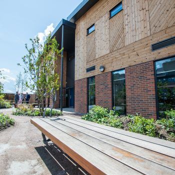 Exterior of a modern building with wood siding, brick, and a wooden bench in a landscaped courtyard.