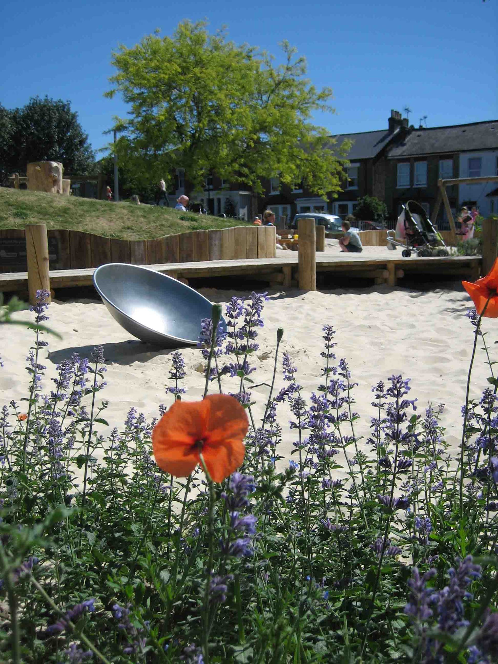 Playground with sand, flowers, a large metal dish, and people on a sunny day.