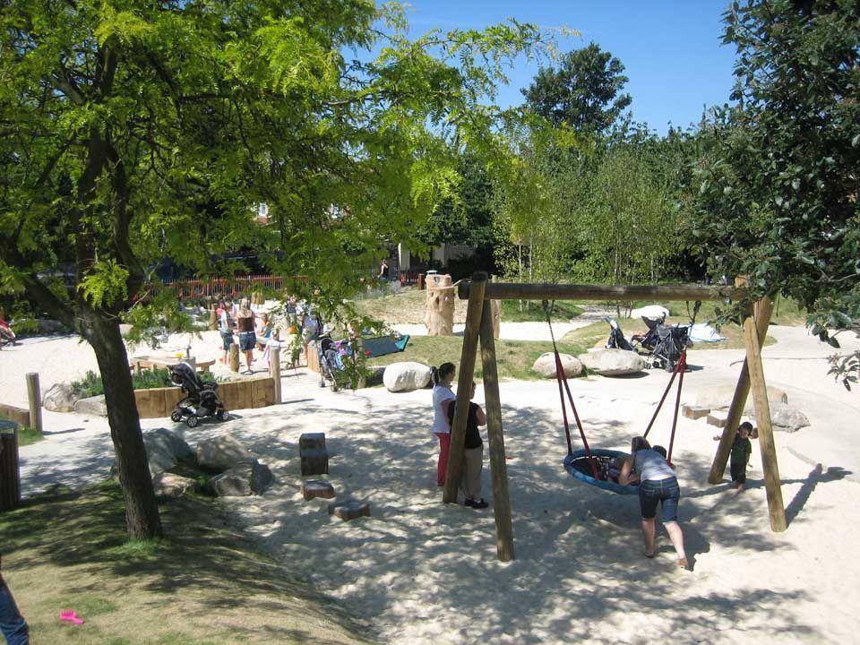 Children playing on swings and a playground with trees and stones, outdoors.
