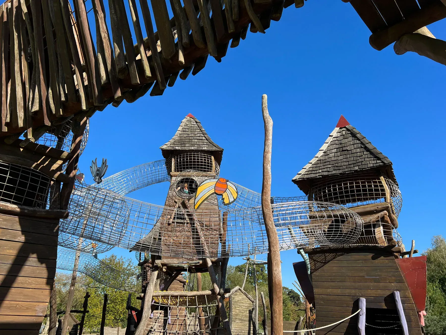 Playground with wooden towers, netting, and bridges against a blue sky.