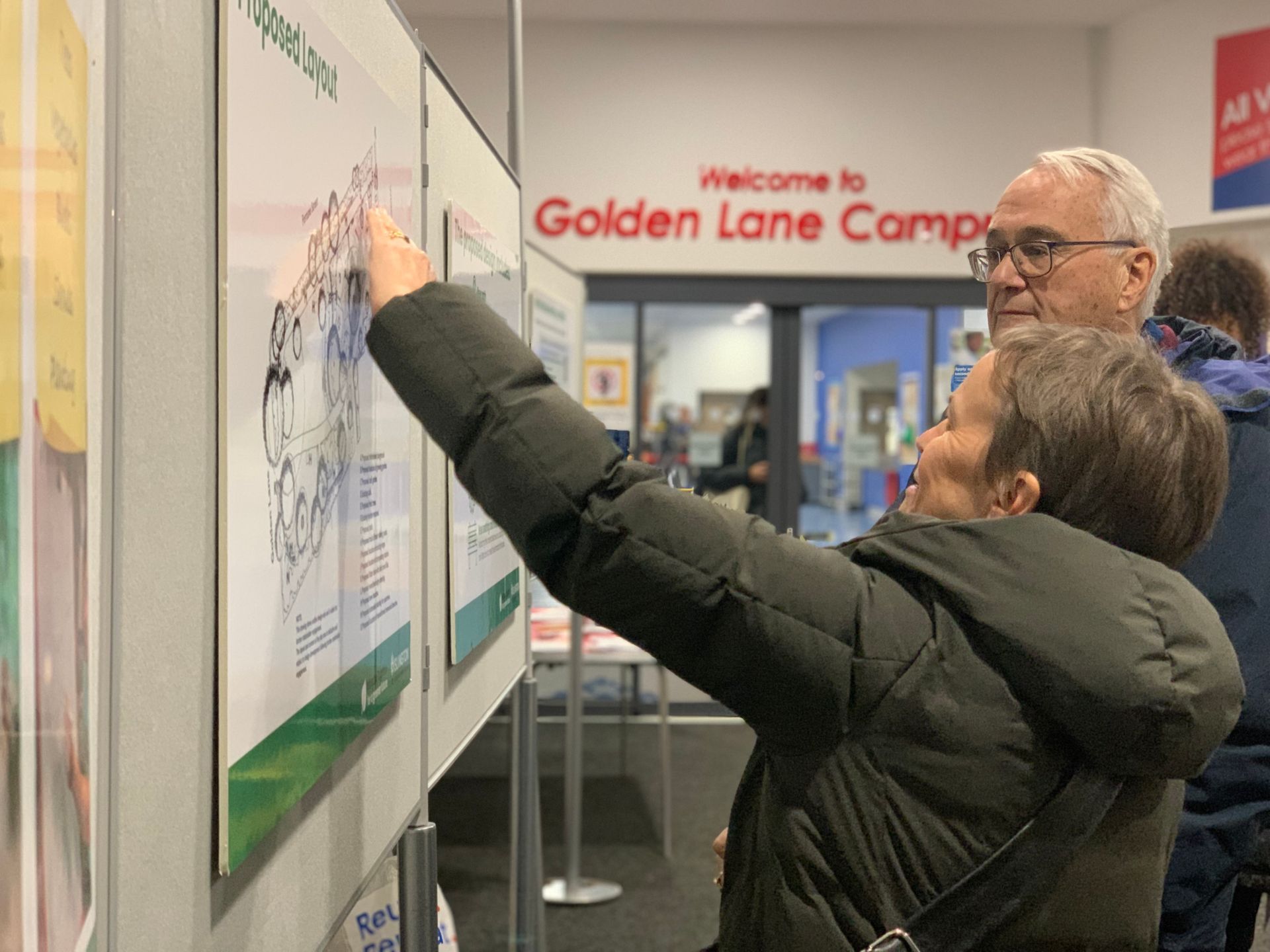 Woman points at map on wall, smiling, with man beside her. Golden Lane Campus sign.