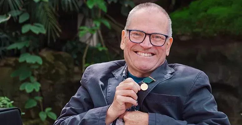Smiling Matthew Biggs in glasses holding a microphone, seated outdoors with lush greenery behind.