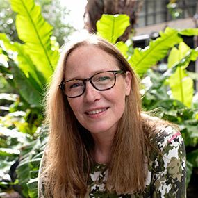Woman with glasses smiles, surrounded by green plants.