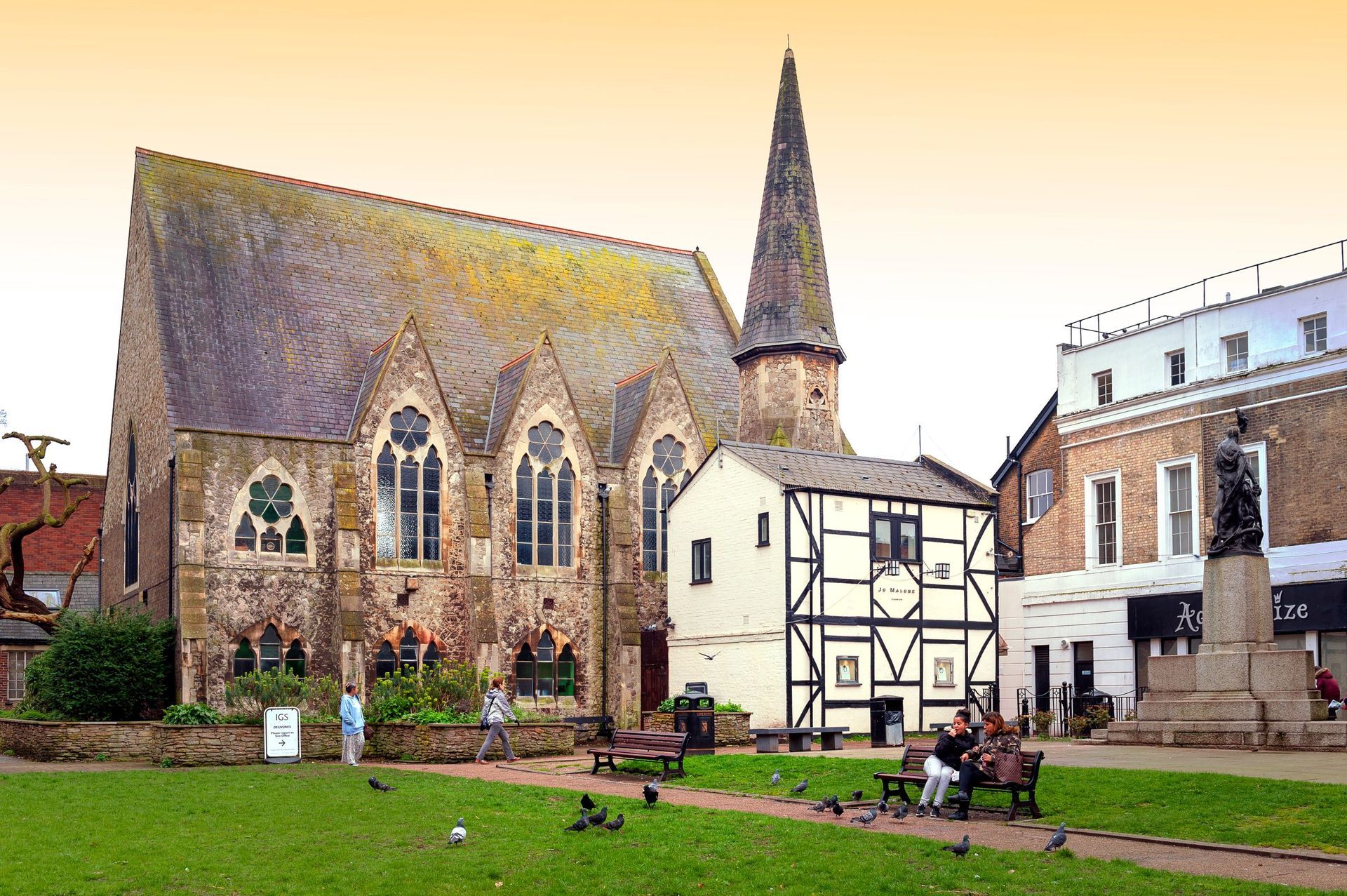 Gothic church and half-timbered building in a town square with statue and people on benches; green lawn.