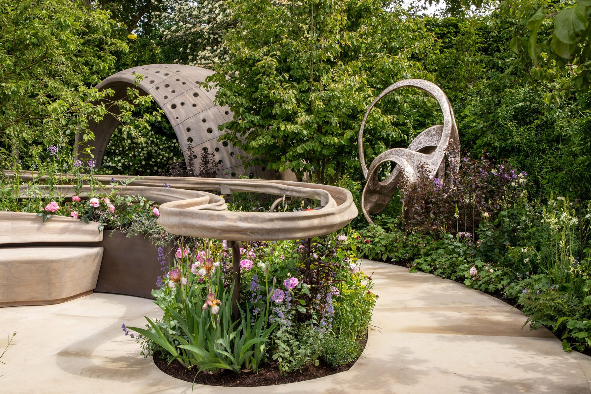 Modern garden with circular stone benches, a perforated metal sculpture, and lush greenery and flowers