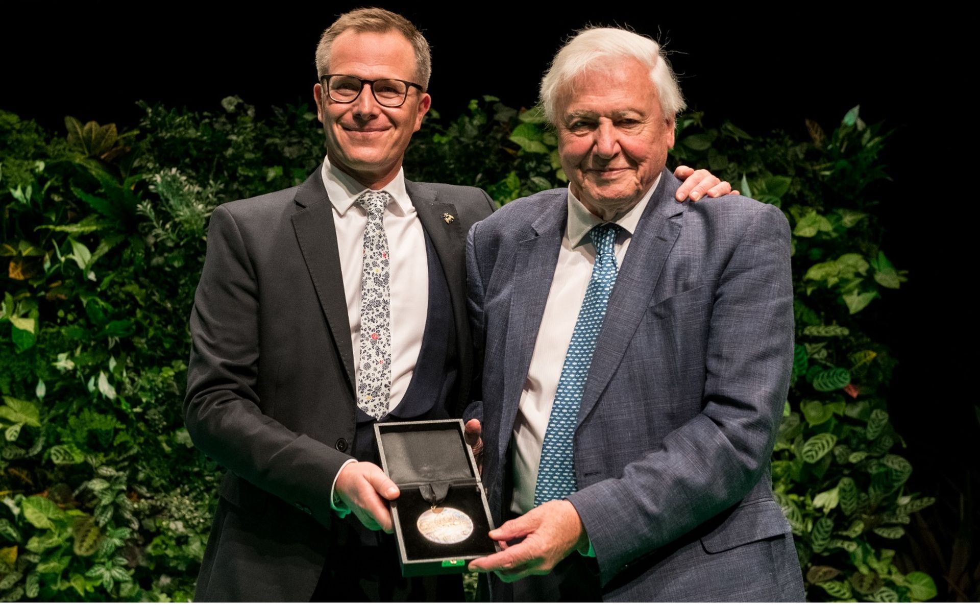 Two men, one in glasses, holding a medal, both smiling. Stage with green backdrop.