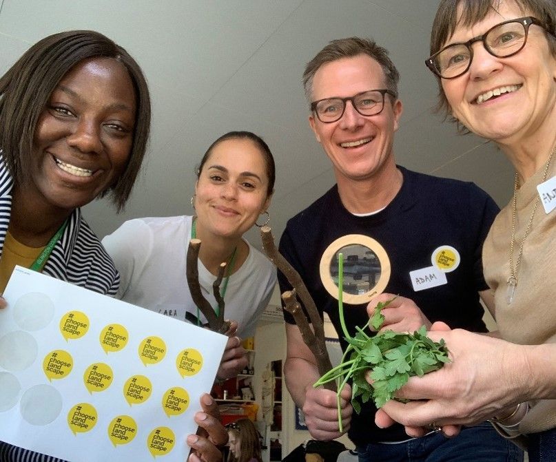 Four smiling people holding stickers, branches, and herbs in a bright room.