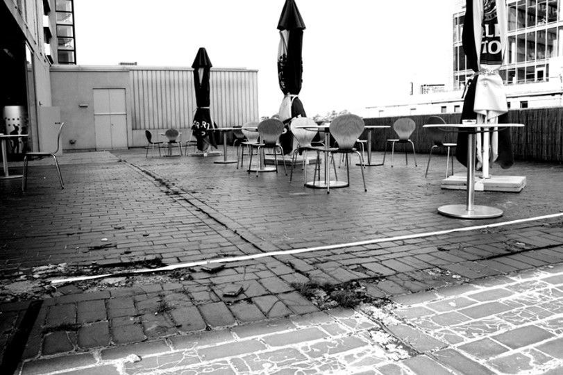 Black and white photo of outdoor patio with tables, chairs, and closed umbrellas on brick.