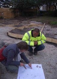 Two people crouch, reviewing a blueprint at a construction site. One wears a high-vis jacket.