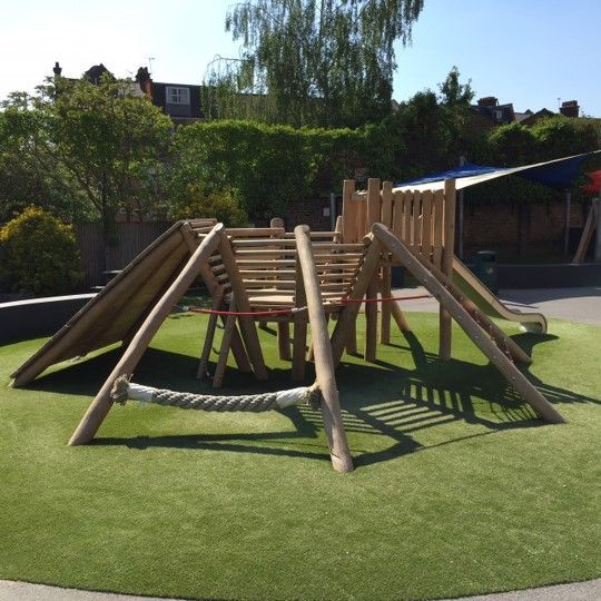 Wooden playground structure on green turf, featuring climbing elements, a slide, and rope bridge.