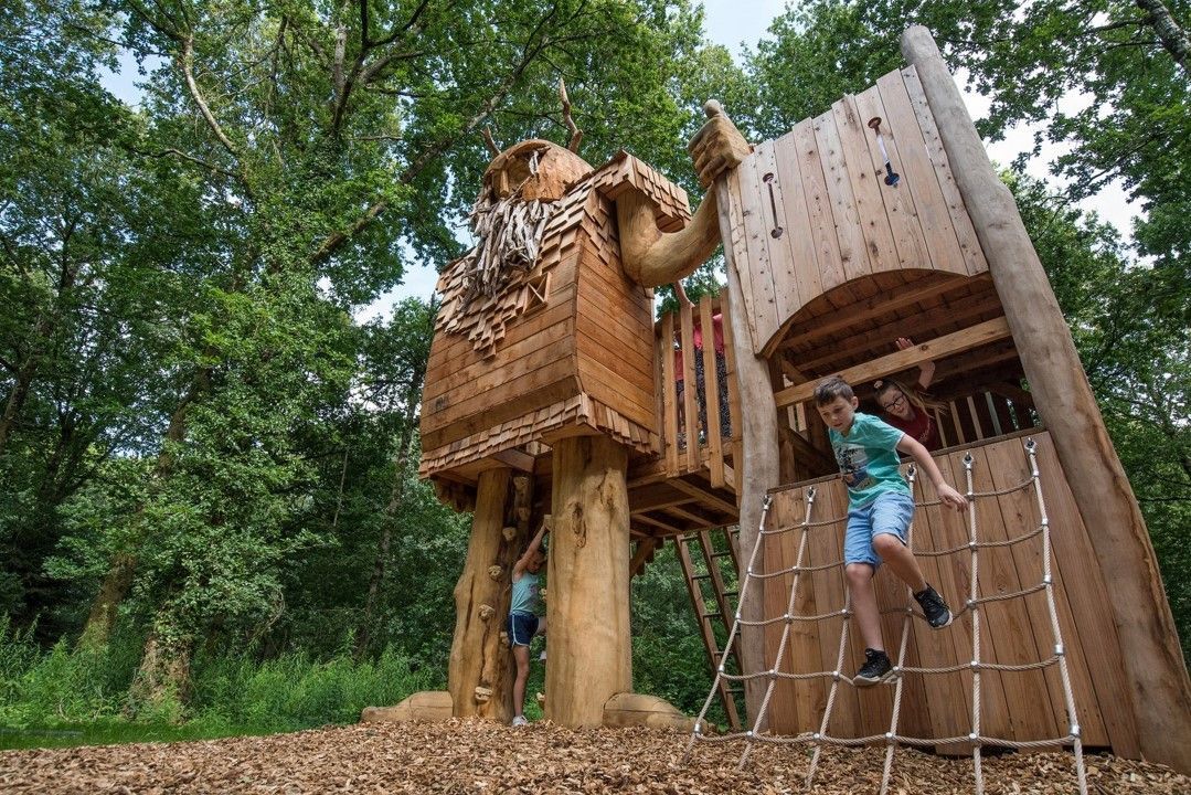 Children playing on a large wooden play structure in a forest.