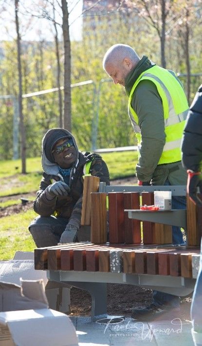 Two men constructing a wooden bench outdoors. One kneels, smiling. The other wears a yellow vest, looking on.