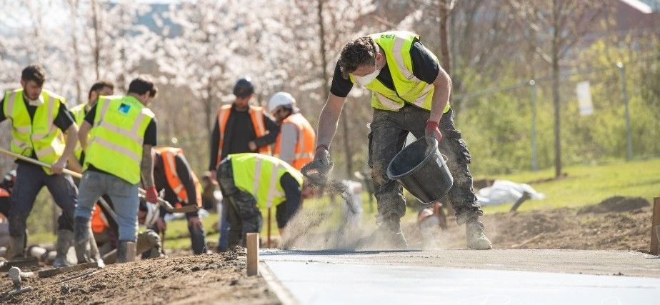 Construction workers wearing vests, laying a road. One is pouring concrete. Outdoors, sunny day.