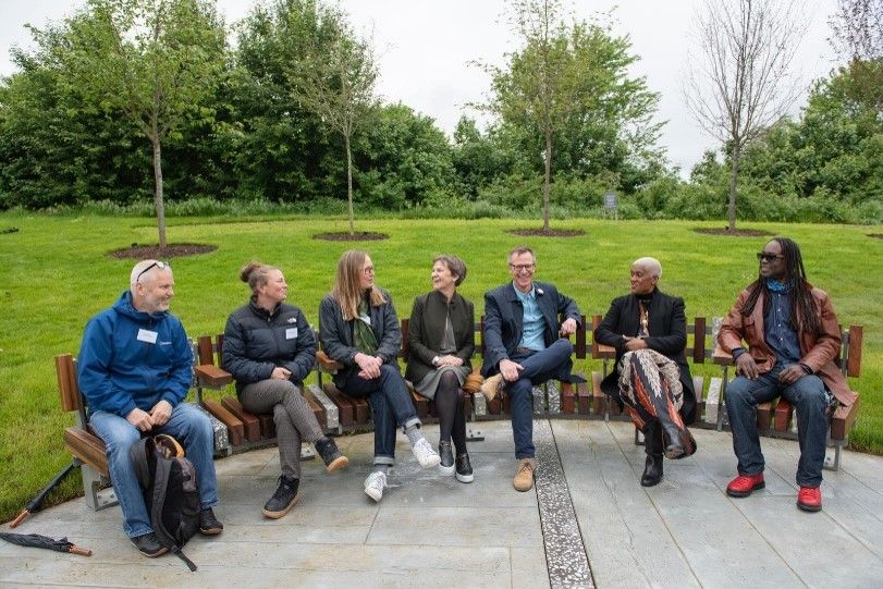 Seven people sit on a bench outdoors, smiling and talking. Trees and green grass in background.