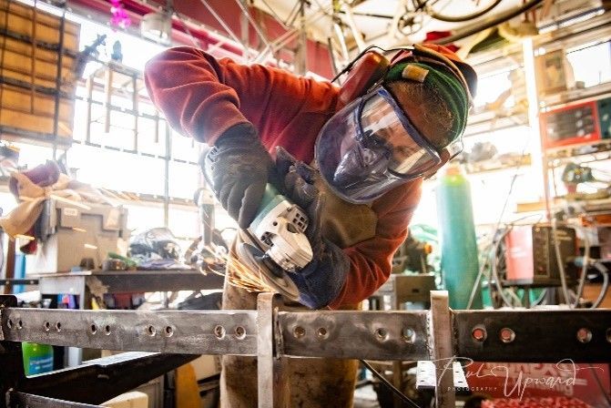 Person grinding metal, wearing protective gear, in a cluttered workshop.