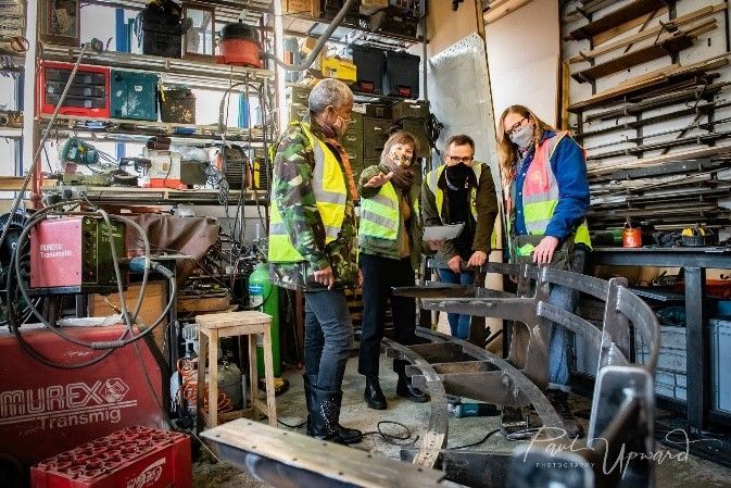 People in safety vests inspect metalwork in a cluttered workshop.