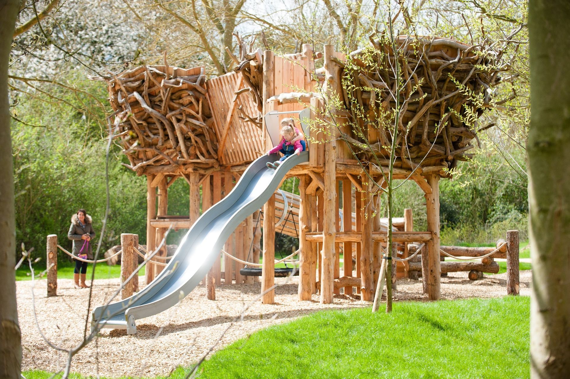 Child sliding down a metal slide on a wooden playground structure, surrounded by trees.