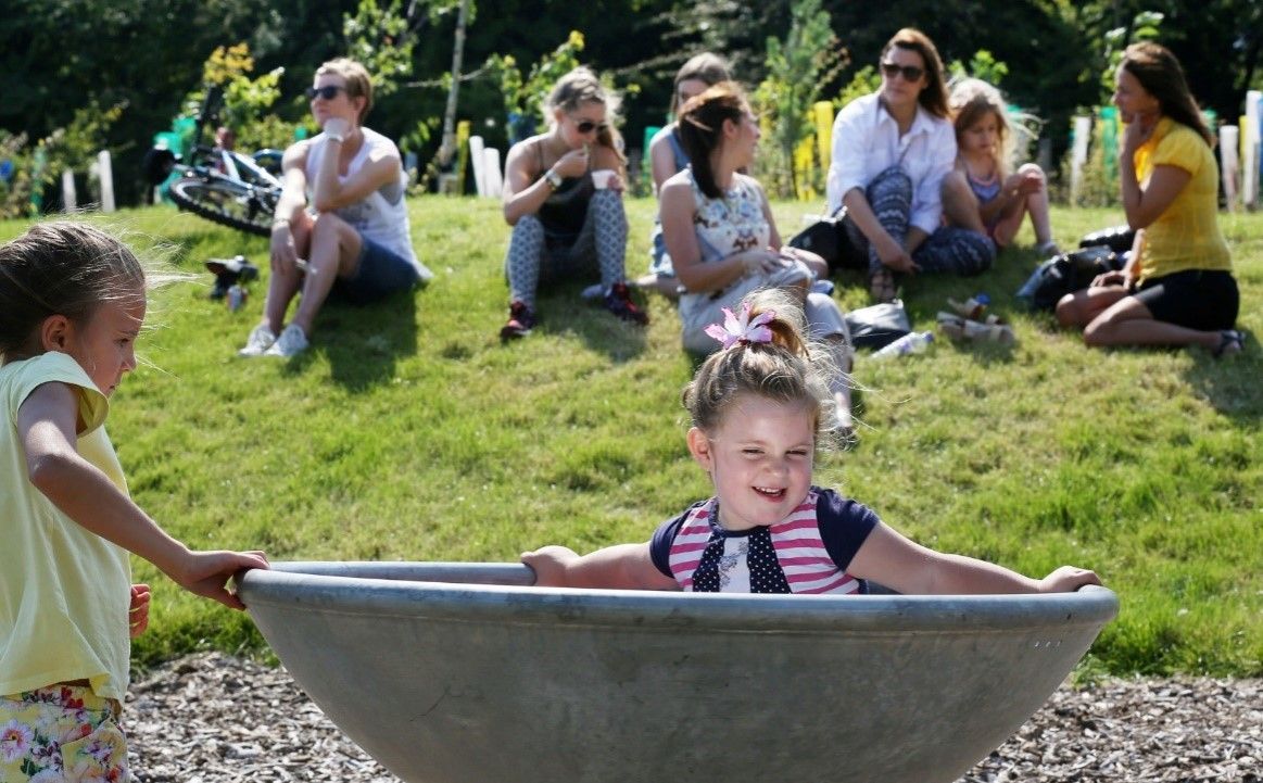Two young girls in a park; one in a bowl-like structure, smiling. Adults watch from a grassy hill.