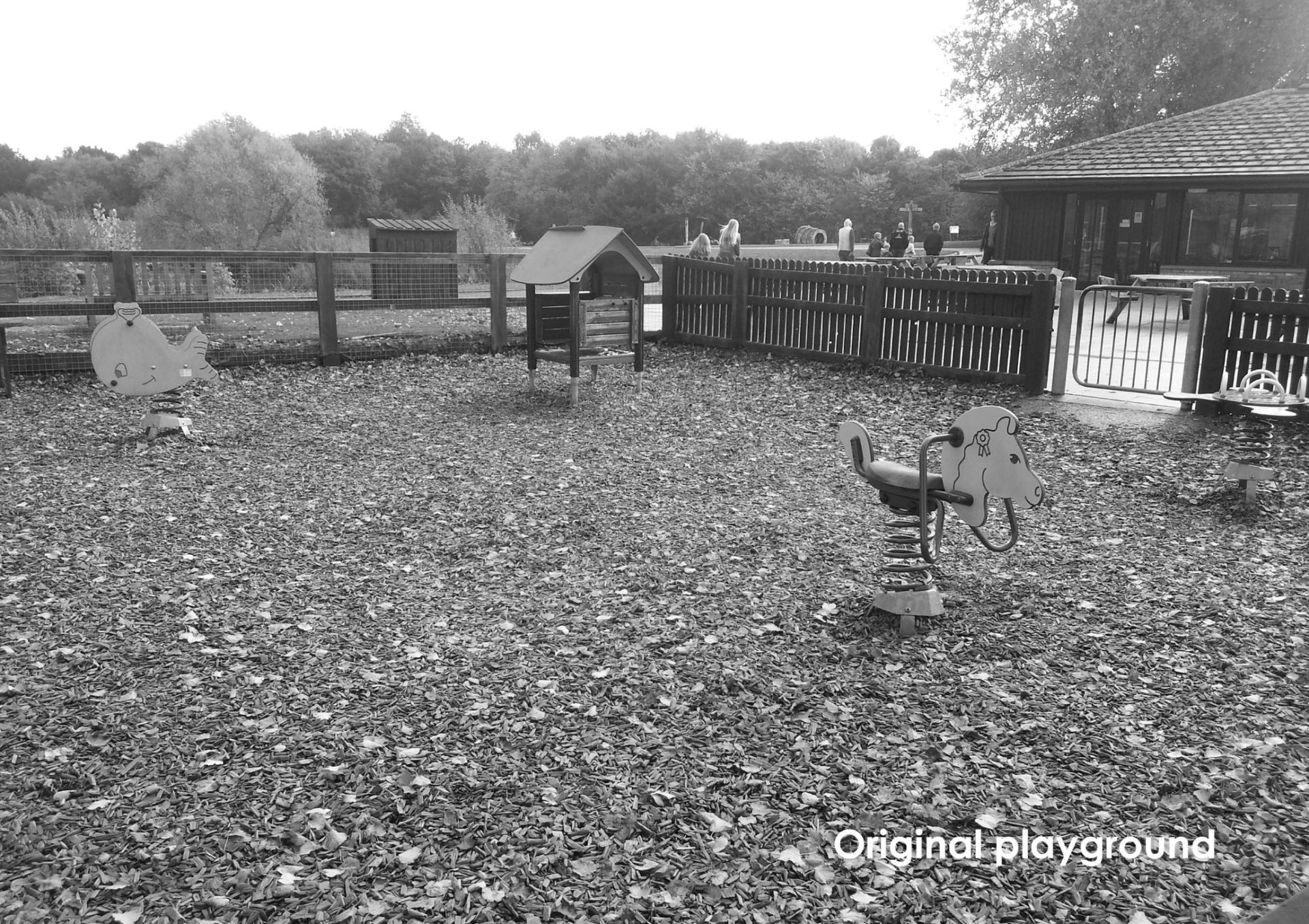 A black and white photo of a playground with several play structures surrounded by a fence and trees.