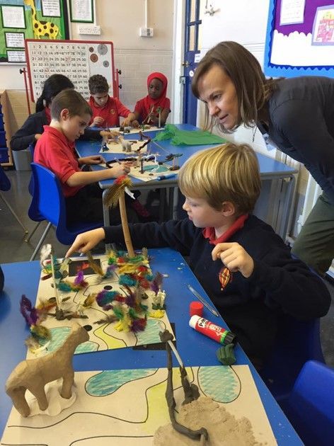A teacher helping a boy create a diorama with colorful decorations and a dinosaur figure in a classroom.