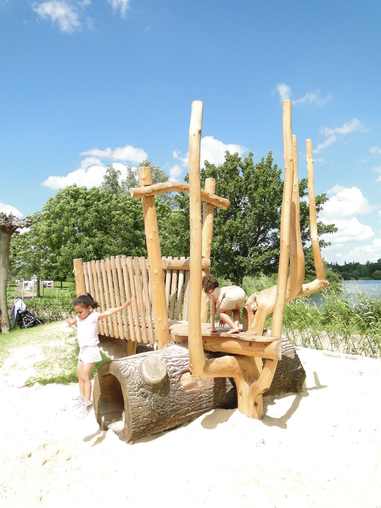 Children playing on a wooden playground structure with a log tunnel, blue sky, and green trees.