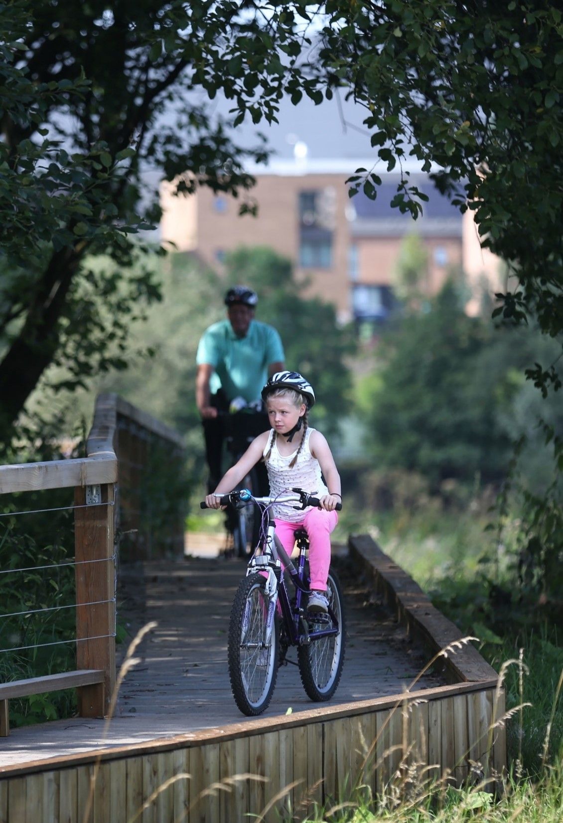 Girl on a bike rides across a wooden bridge, followed by an adult, with a building in the background.