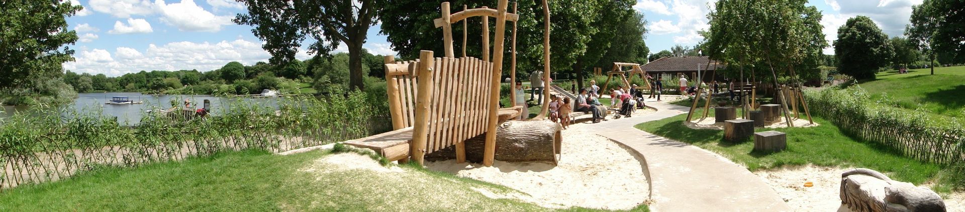 A park with wooden playground equipment, people, grass, trees, and a lake under a blue sky.