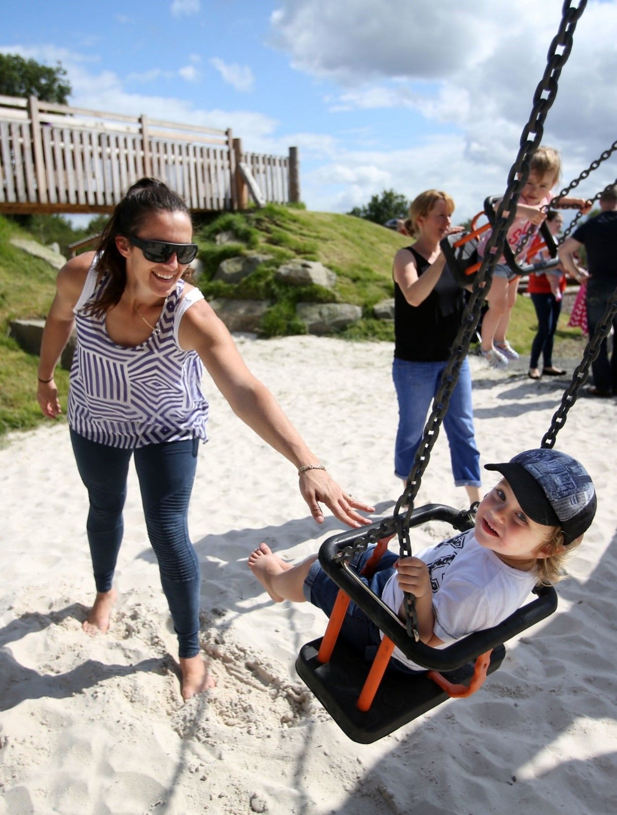 Woman pushes child on a swing at a playground. Bright sunny day. Other people in background.