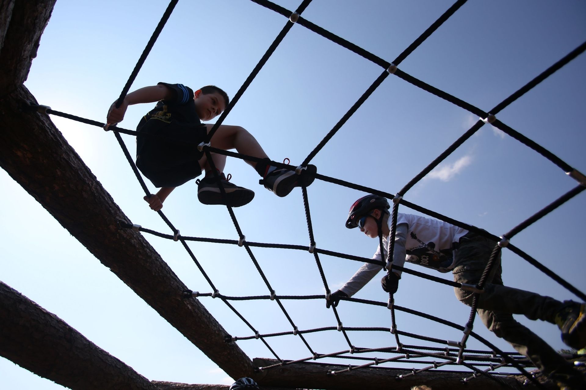 Two people climbing a black rope playground structure against a bright sky.