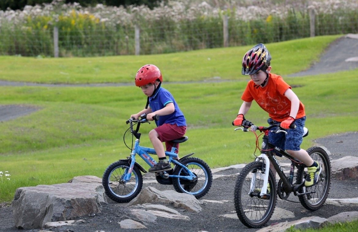 Two young boys on bikes ride a pump track, wearing helmets, on a green field.