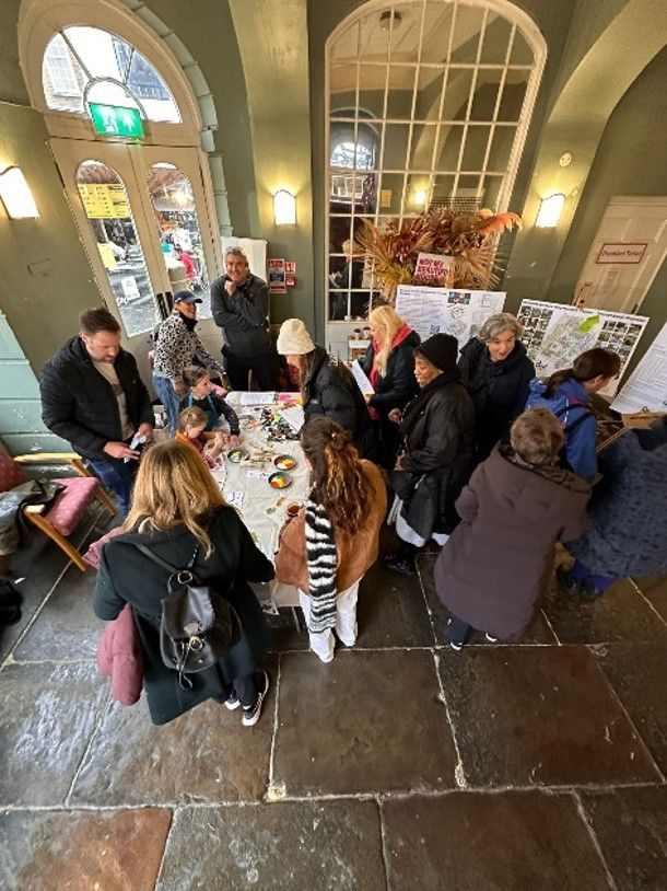 People browse a display table with goods in a hall. Others chat, indoors, natural light.