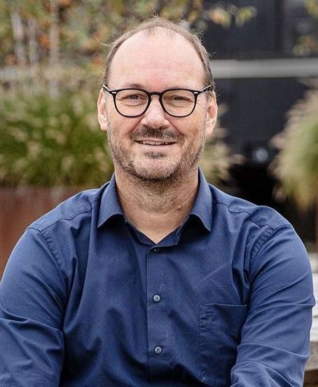 Man with glasses, smiling, wearing blue shirt, outdoors, blurred background.