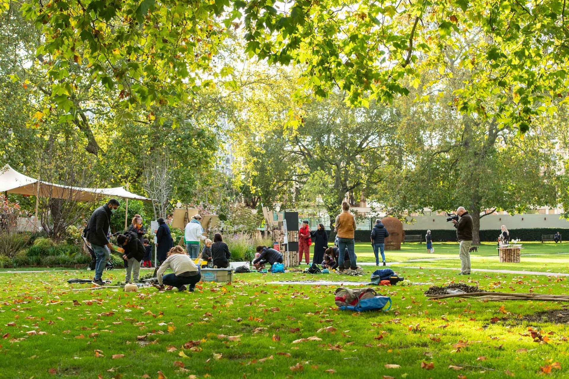 People setting up equipment in a sunny park with fallen leaves. Trees, tents, and art are in the background.