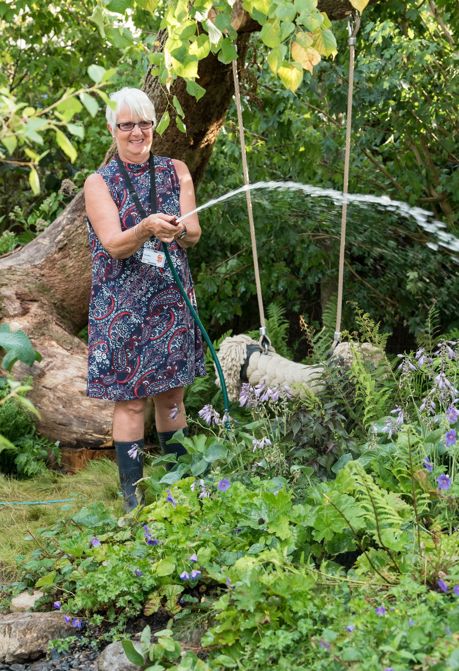 Woman in a colorful dress waters plants in a lush garden, swing hanging from a tree.