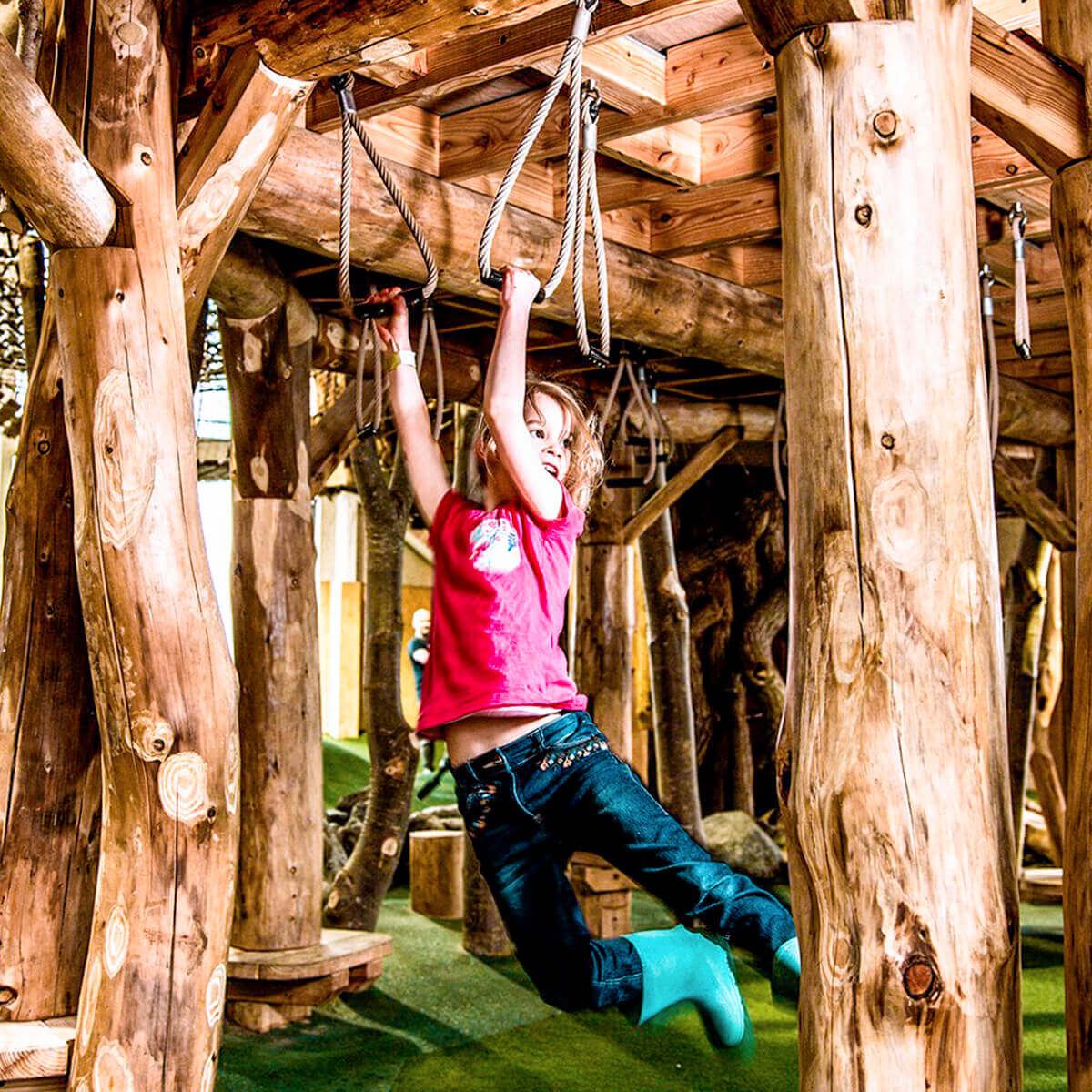 Girl in pink shirt hangs from wooden monkey bars at indoor playground.