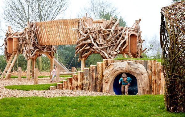 Children playing in a wooden playground with tunnels, bridges, and climbing structures outdoors on a grassy field.