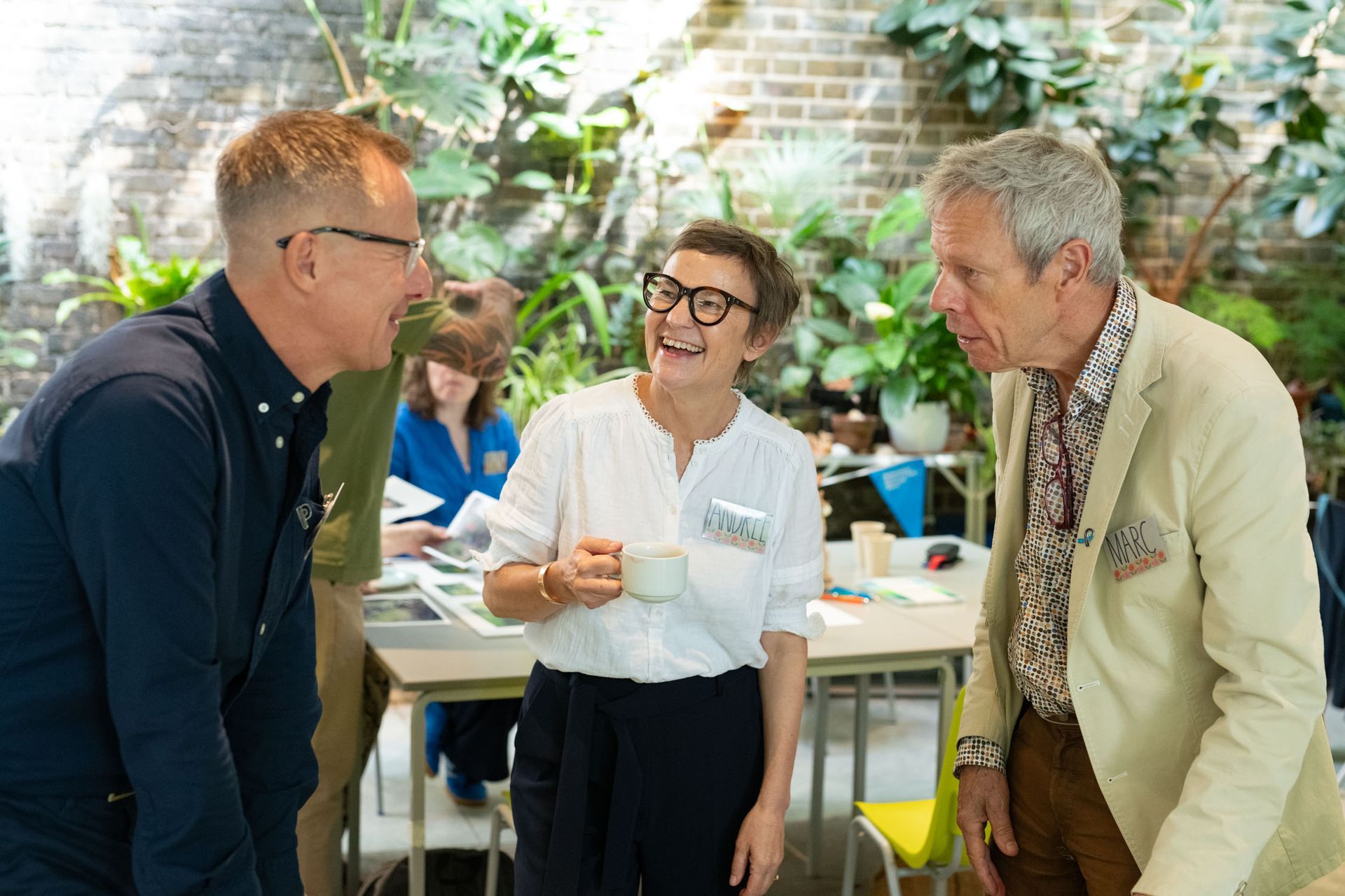 Three people chatting at a garden event, with greenery and tables in the background.