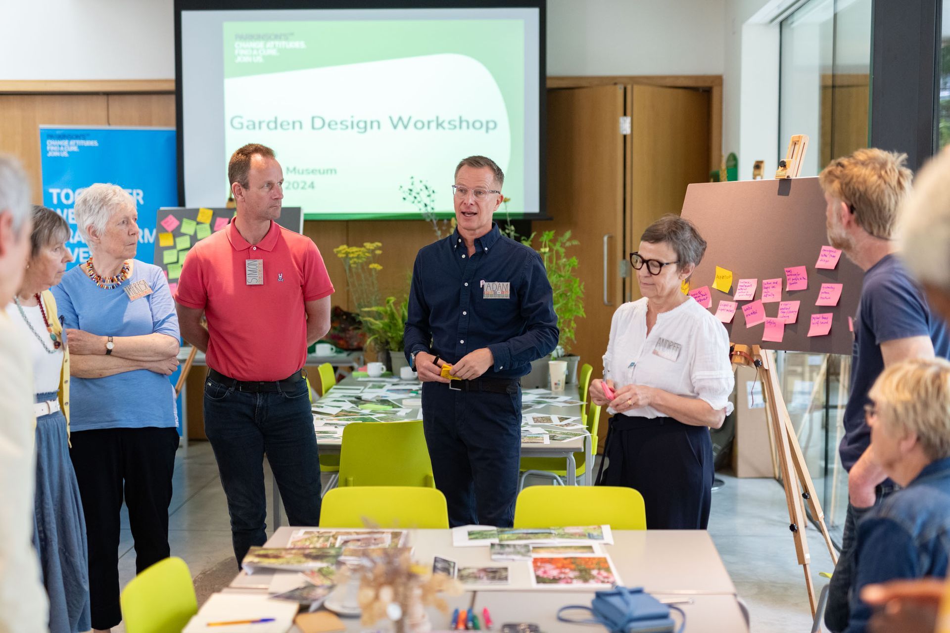 Workshop participants standing around a table in a bright meeting room with a presentation screen and sticky notes