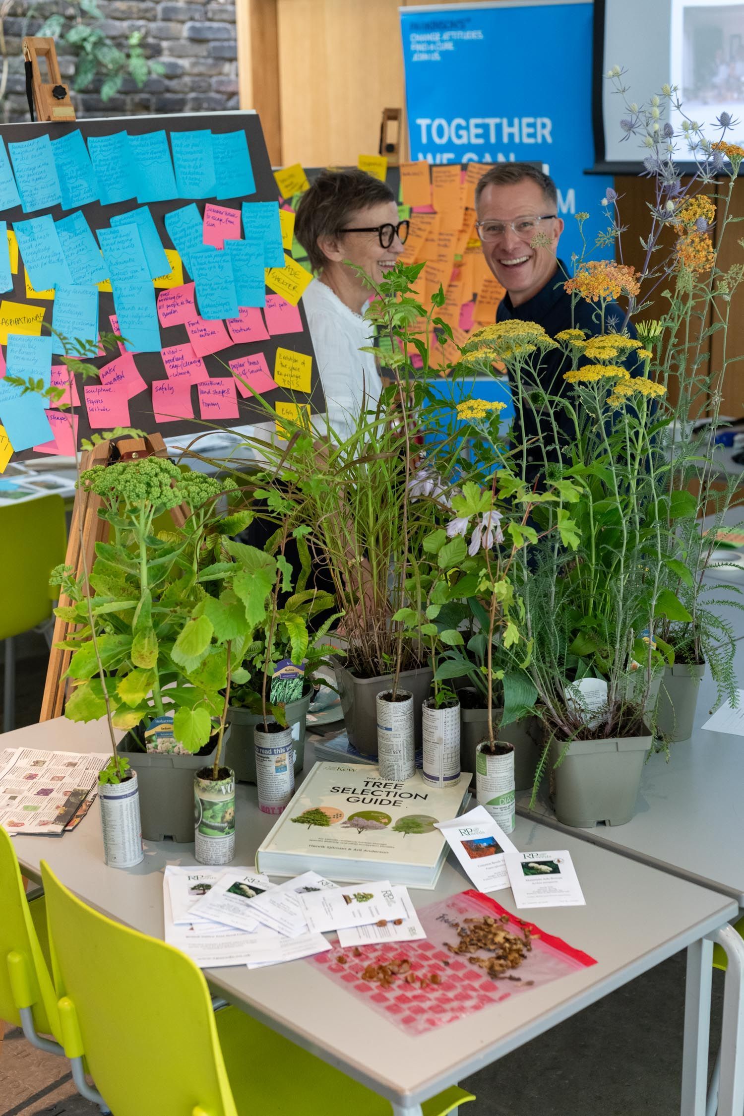 Two people behind a plant display with colorful sticky notes and a blue “TOGETHER” sign in a workshop room
