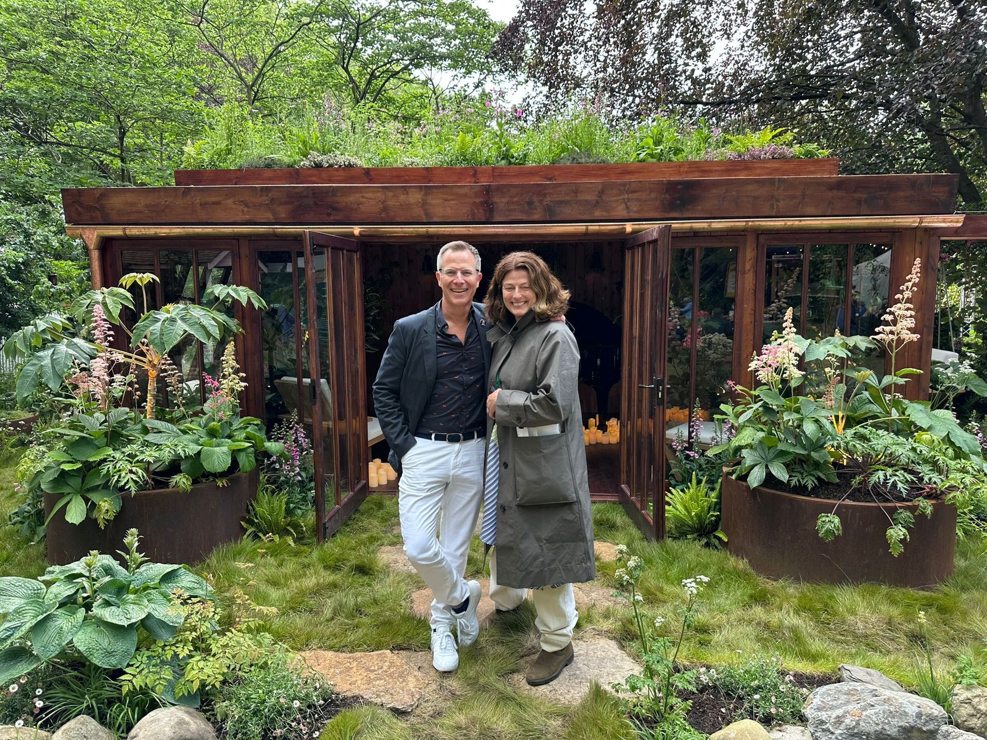 Two people posing in a lush garden under a wooden arbor, surrounded by greenery and potted plants.