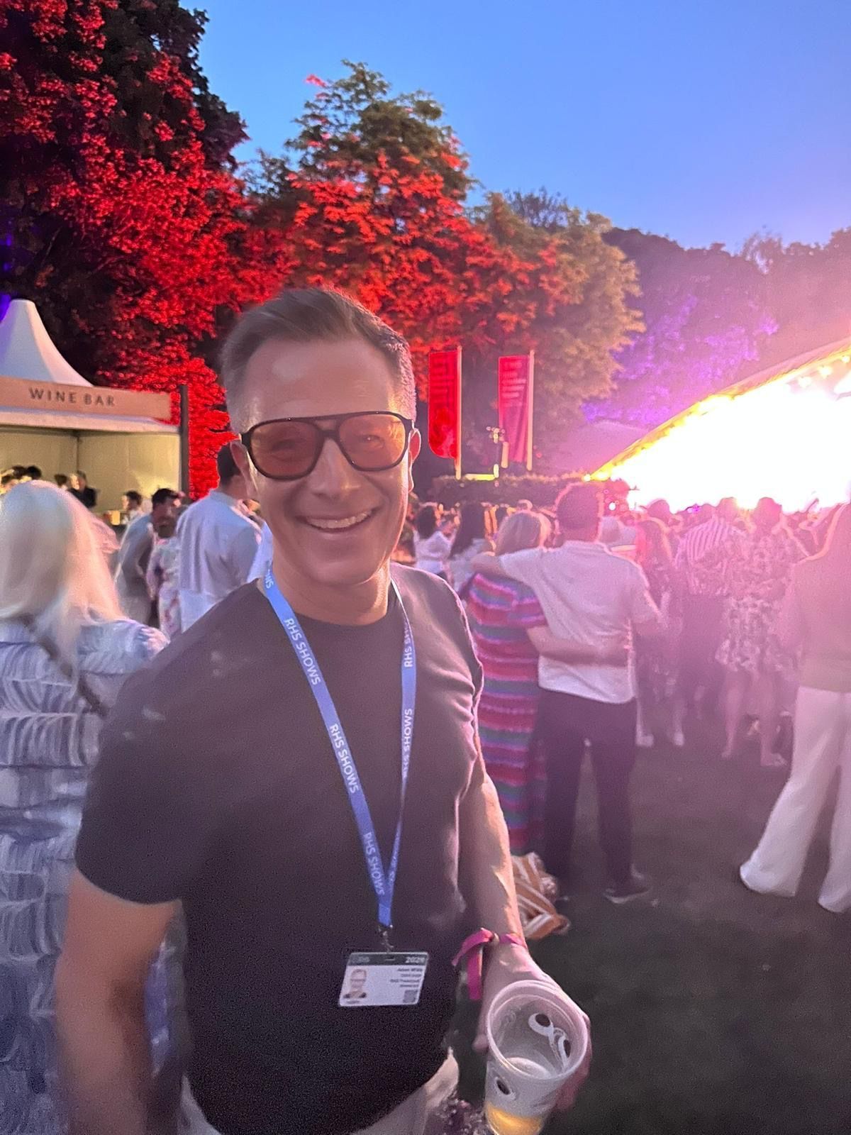 Smiling Adam White  with Jo Whiley and lanyard at an outdoor event with bright sunset and crowd