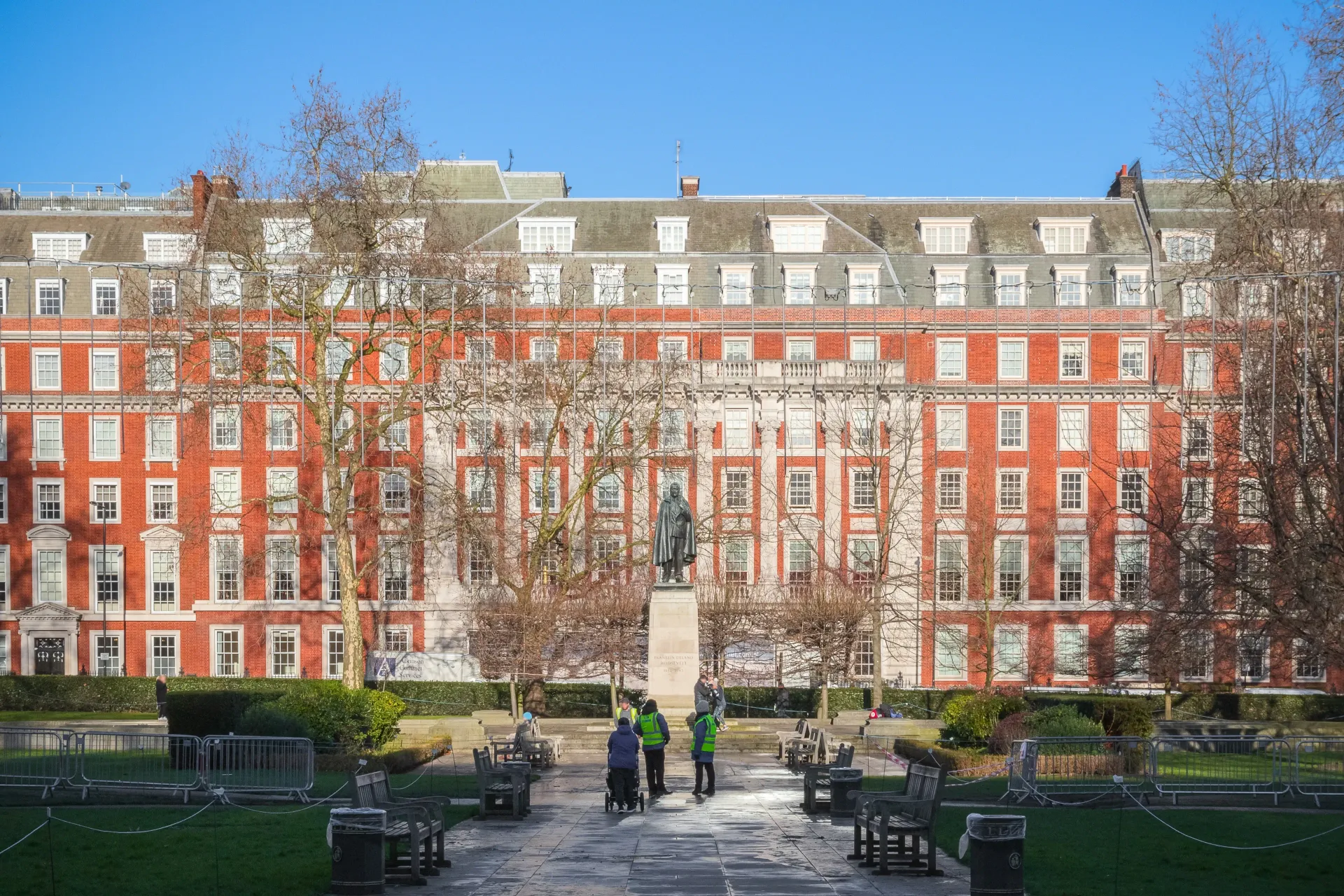 Red brick building, statue, and people in a park. The sky is blue and the trees are bare.