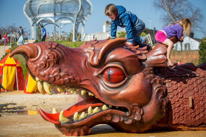 Two children climbing a red dragon sculpture's head outdoors, with a gazebo and tents in the background.
