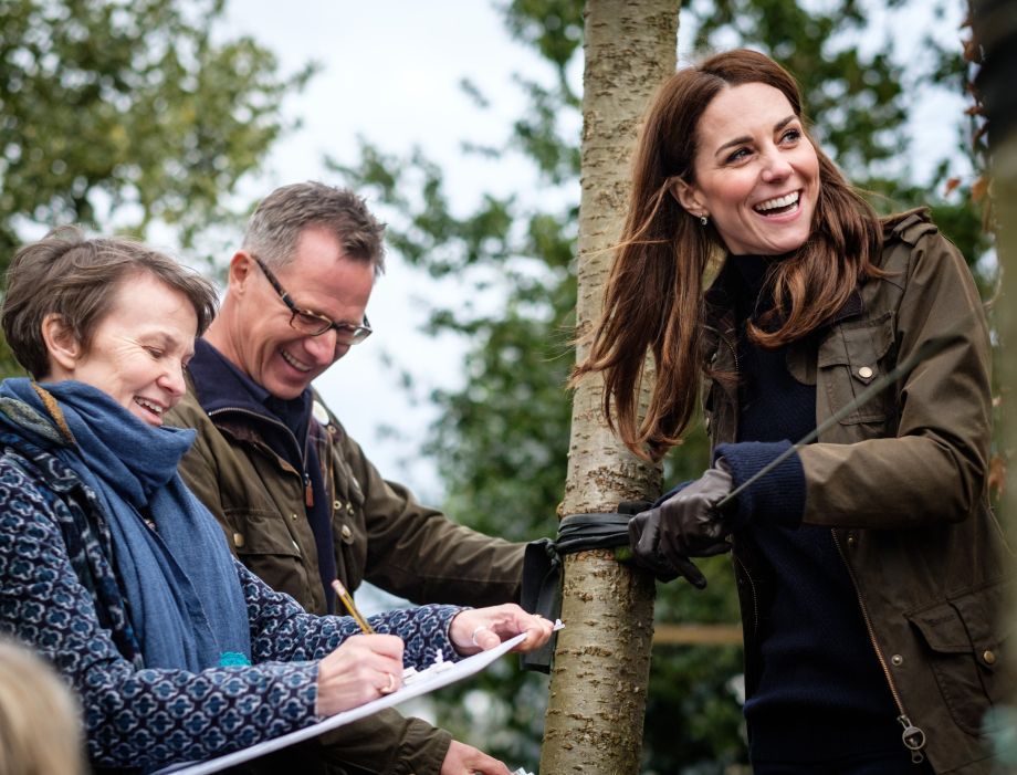 Kate Middleton smiles planting a tree. Two people look on, taking notes. Outdoors, daytime.