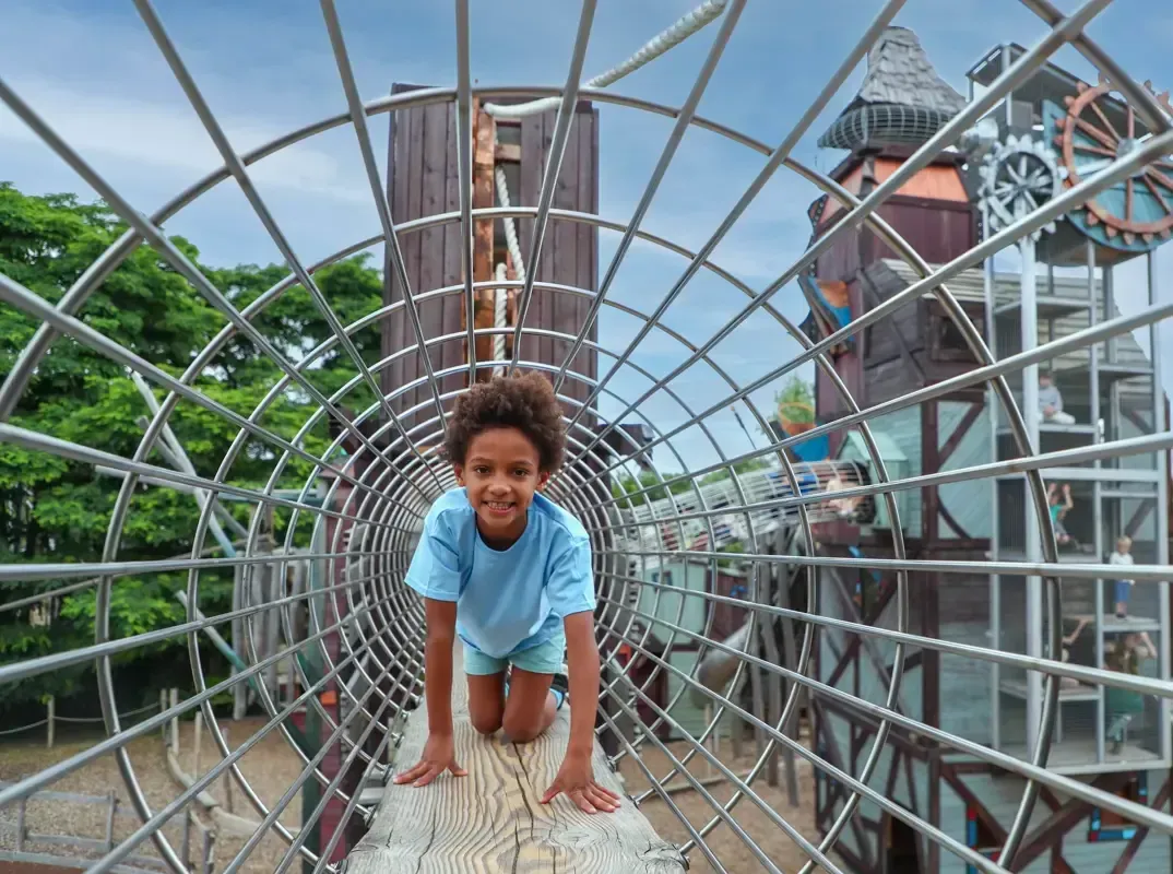 Young child smiles while crawling through a metal tunnel at a playground.